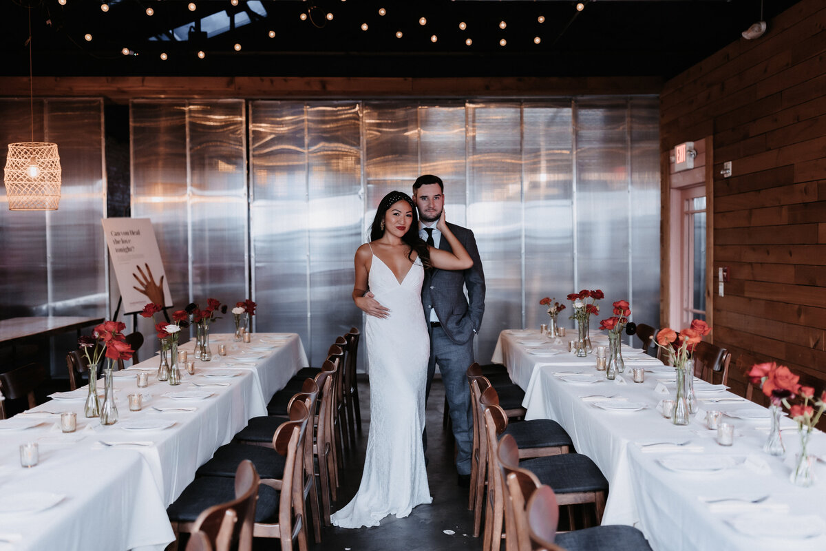 Bride and groom posing between long tables with ivory tablecloths in an industrial style space