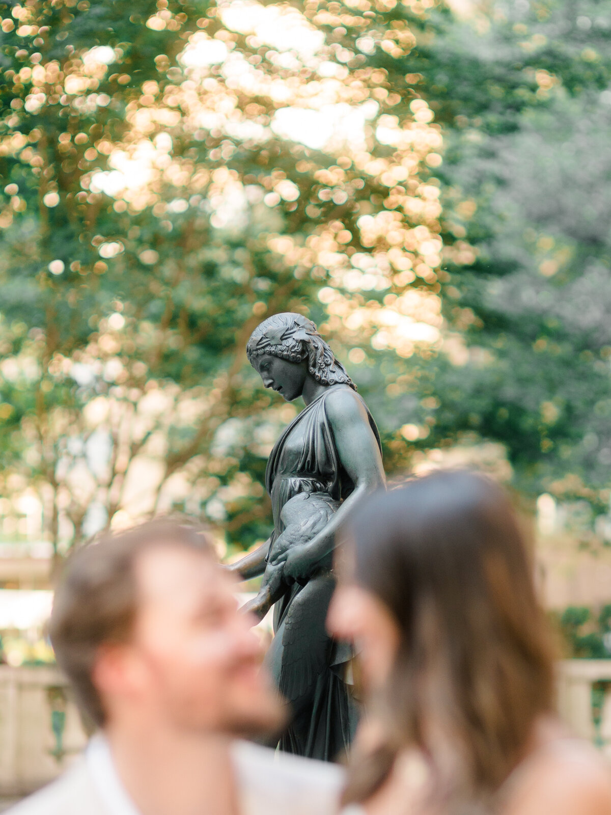 A bronze statue of a woman holding a bird stands in soft focus behind a couple sharing a quiet moment in Rittenhouse Square. Dappled golden light filters through the trees, adding a romantic, timeless ambiance. Captured by Elizabeth Kane Photography, a Luxury Philadelphia Wedding Photographer.
