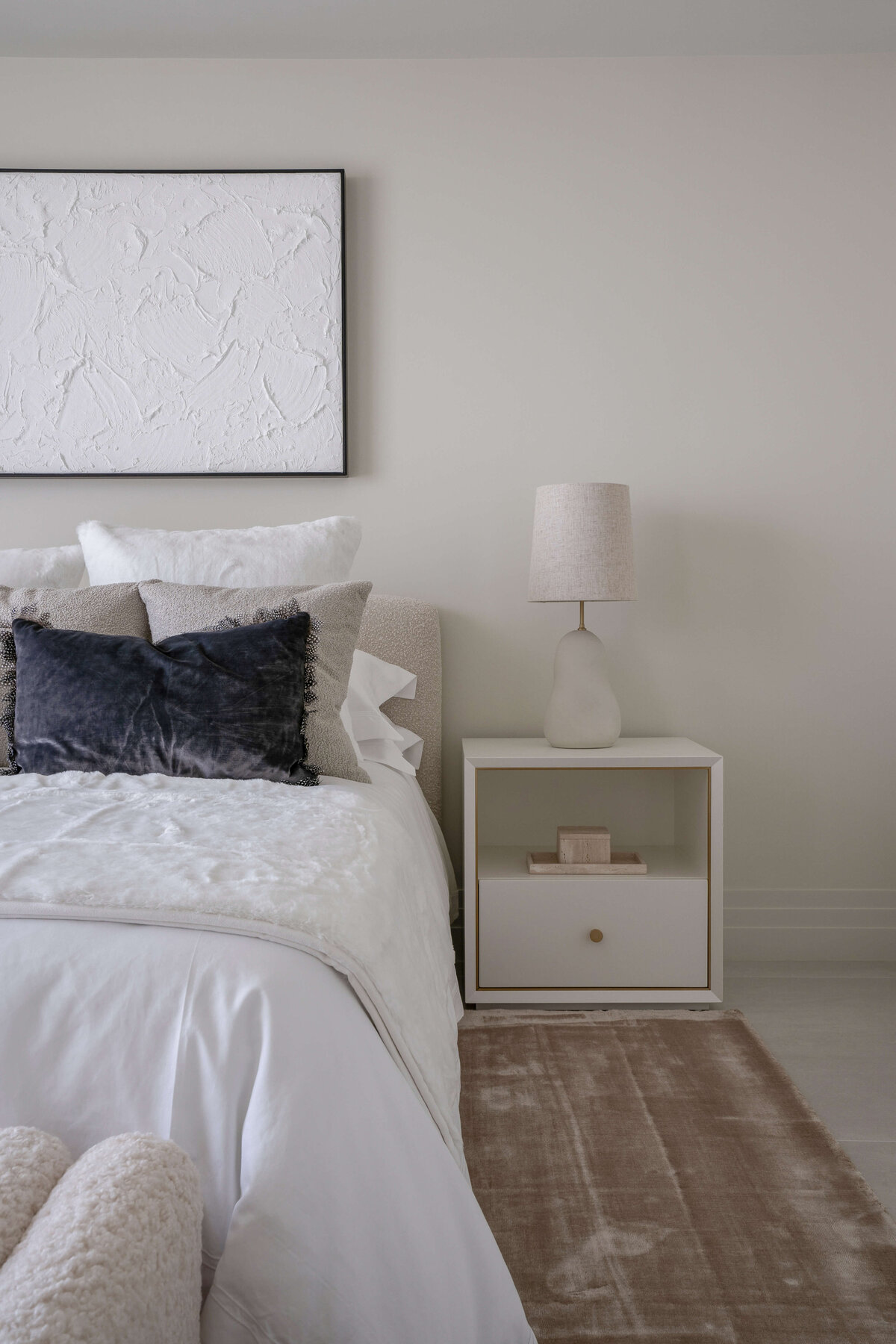 Light and airy guest room featuring a white bed, cream colored bench, and white side tables atop a soft tan rug. Above the bed is a white art piece.