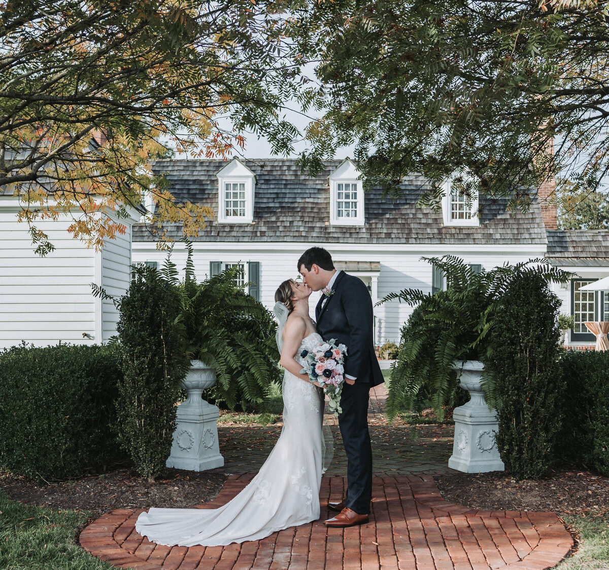 Newlywed couple sharing a kiss in front of the charming Burlington Weddings and Events venue, surrounded by lush greenery and ferns. The bride is holding a bouquet of flowers, and the groom is in a navy suit. Photo by Quinn Photography LLC.