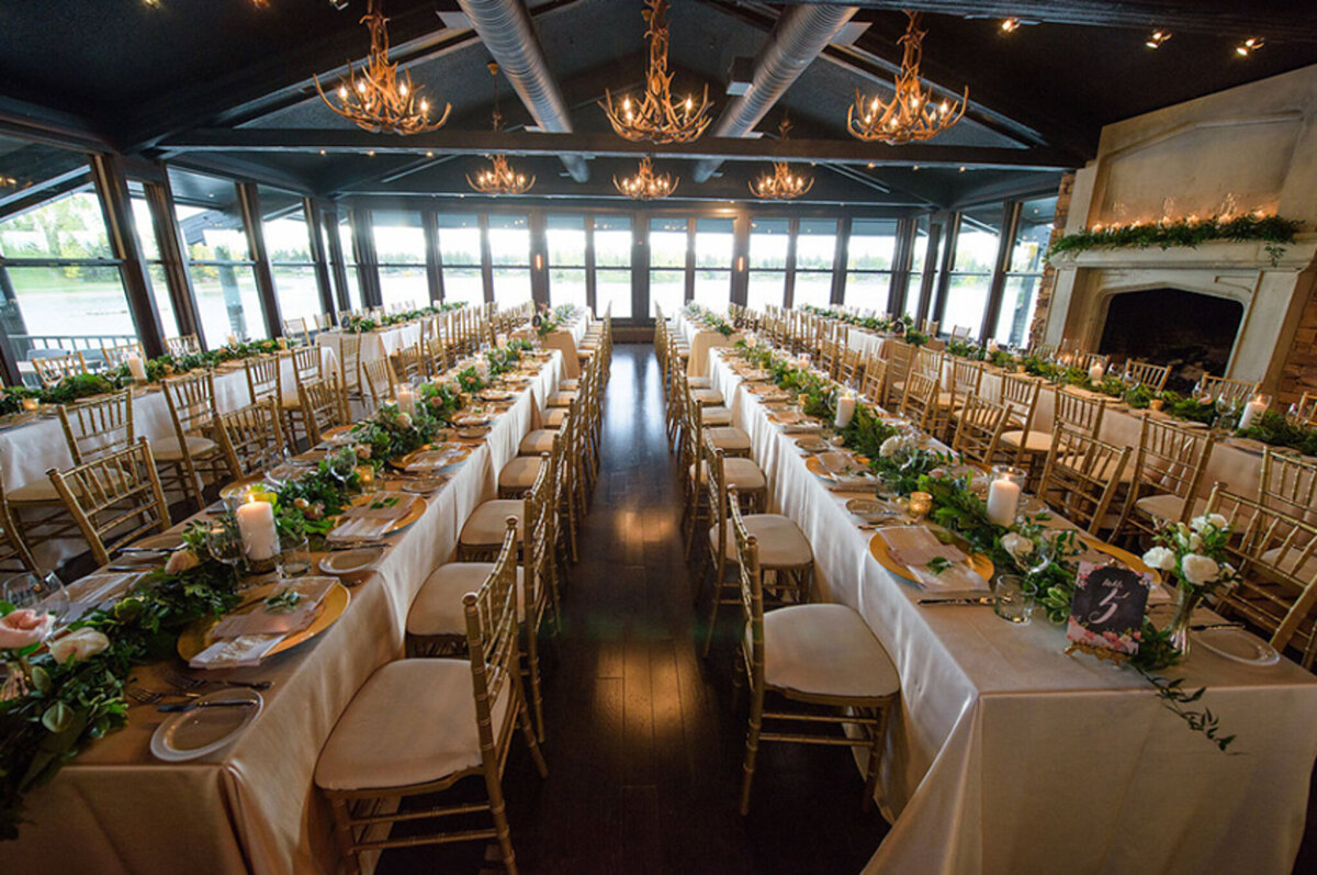 A warm and intimate wedding reception at The Lake House in Calgary, viewed from outside through large glass windows. Guests are seated at long, candlelit tables, engaged in conversation and enjoying the elegant atmosphere. The rustic charm of the venue is highlighted by stone walls, a grand fireplace adorned with greenery and candles, and antler chandeliers casting a soft glow. The dim lighting and reflections in the glass add depth to the scene, creating a cozy and romantic ambiance for the celebration.