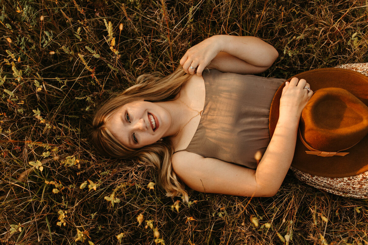 High school senior laying down in the grass in a field and holding a hat