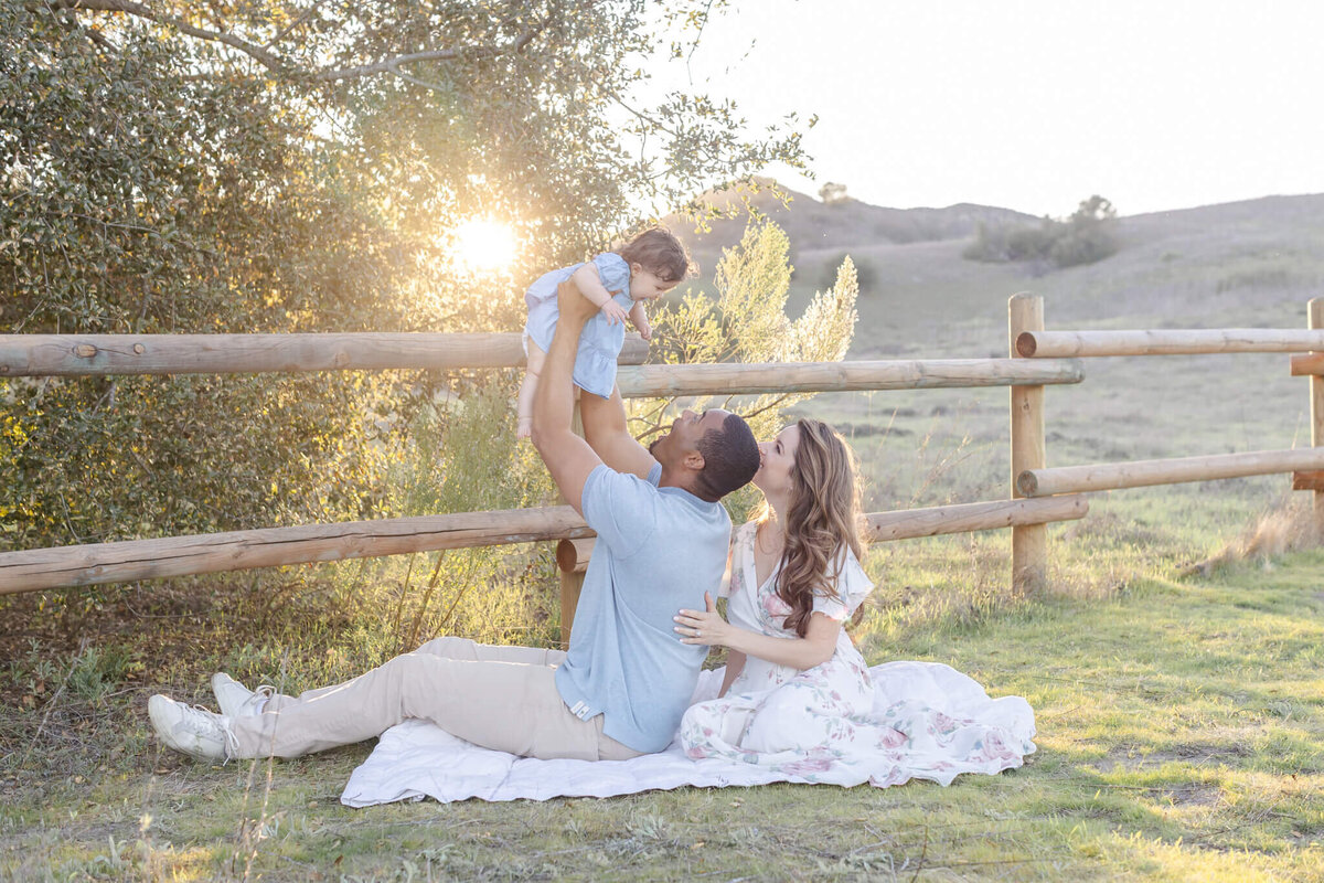 mom and dad on a blanket near a fence and trees with the dad holding the toddler up to the sky
