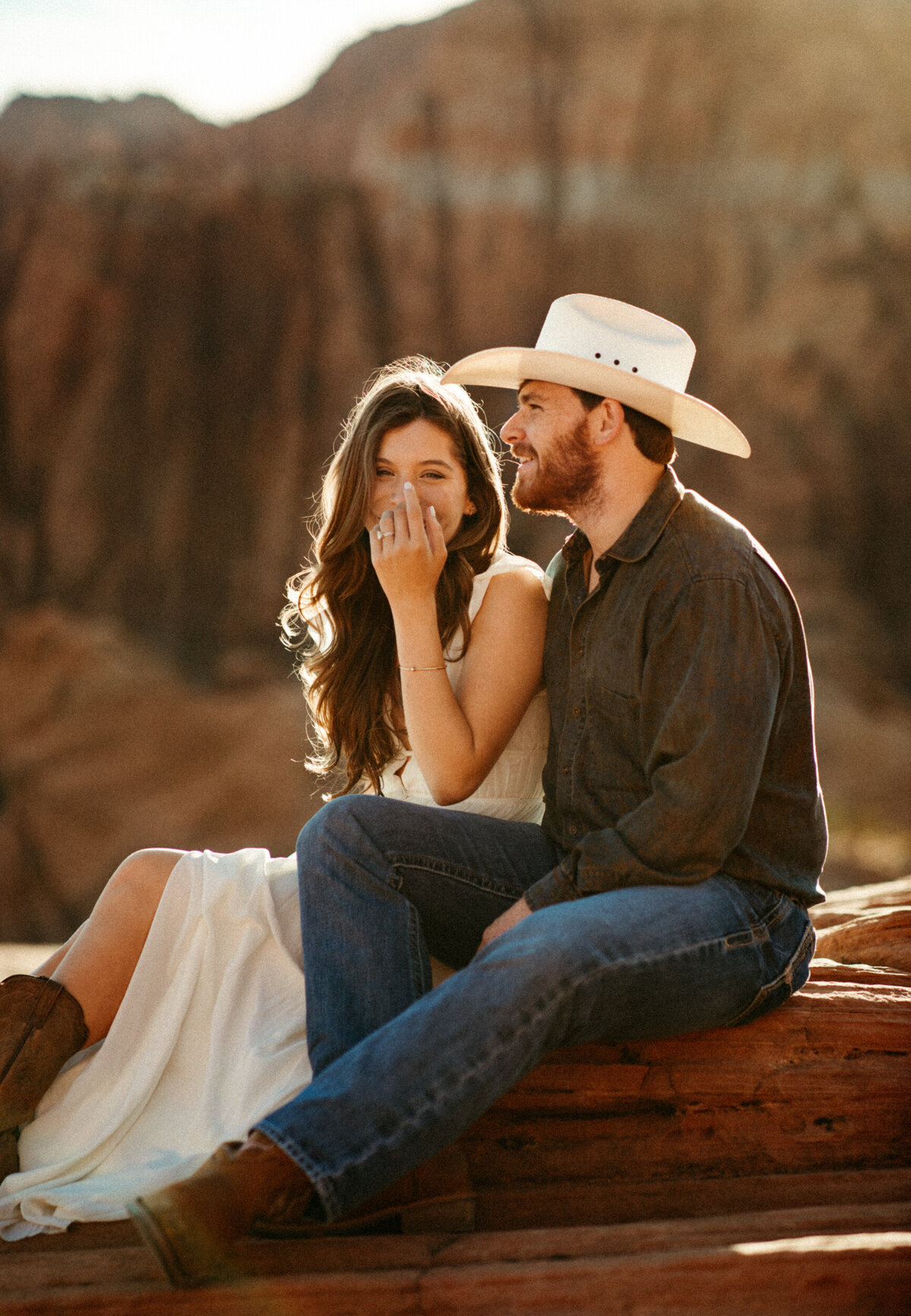 Girl in long white dress and cowboy boots laughing as her husband whispers in her ear in the desert