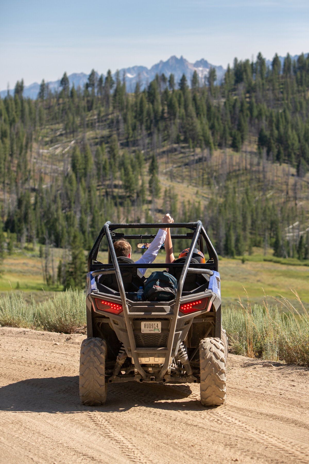 A side-by-side is parked on a dirt road with green pine trees in front of it and mountains all around. The couple in the side-by-side are holding hands and have them raised in the air.