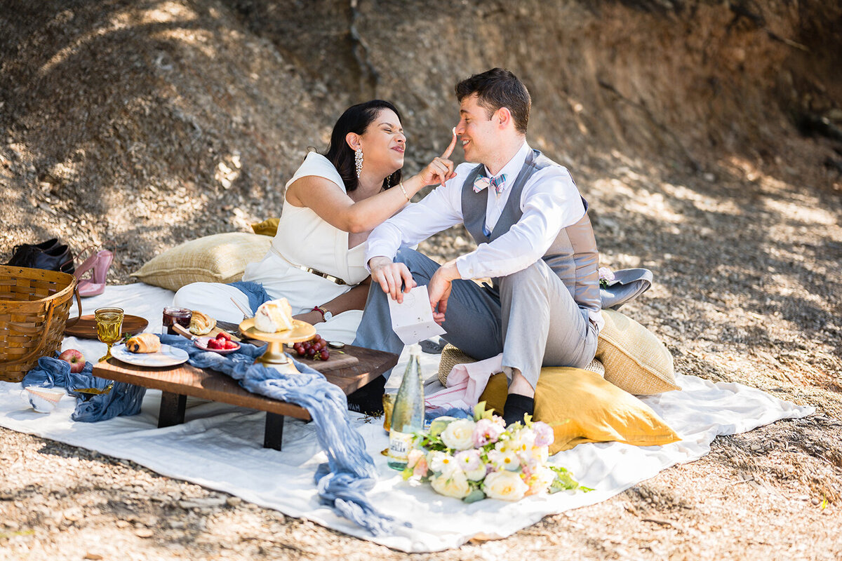 A couple sits down for a brunch picnic on their elopement day along the shores of Carvin’s Cove in Roanoke, Virginia. The picnic setup includes a picnic basket, a couple of pillows, a small table with a draping blue fabric, and all of their food, including croissants, various fruits, cakes, jam, and honey. One marrier takes a bit of frosting on her finger and places it on her partner’s nose in a silly manner.