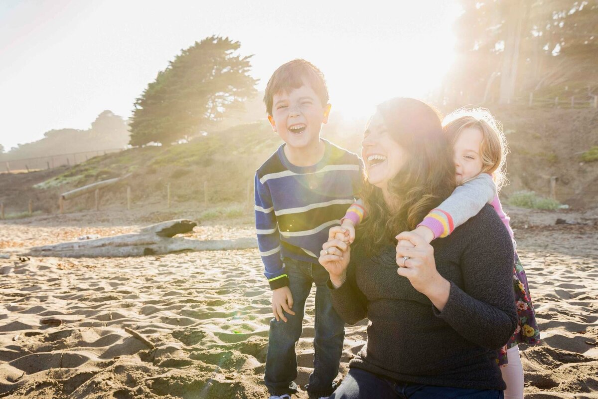 mother, son, and young daughter laughing and hugging at baker beach in san francisoc.