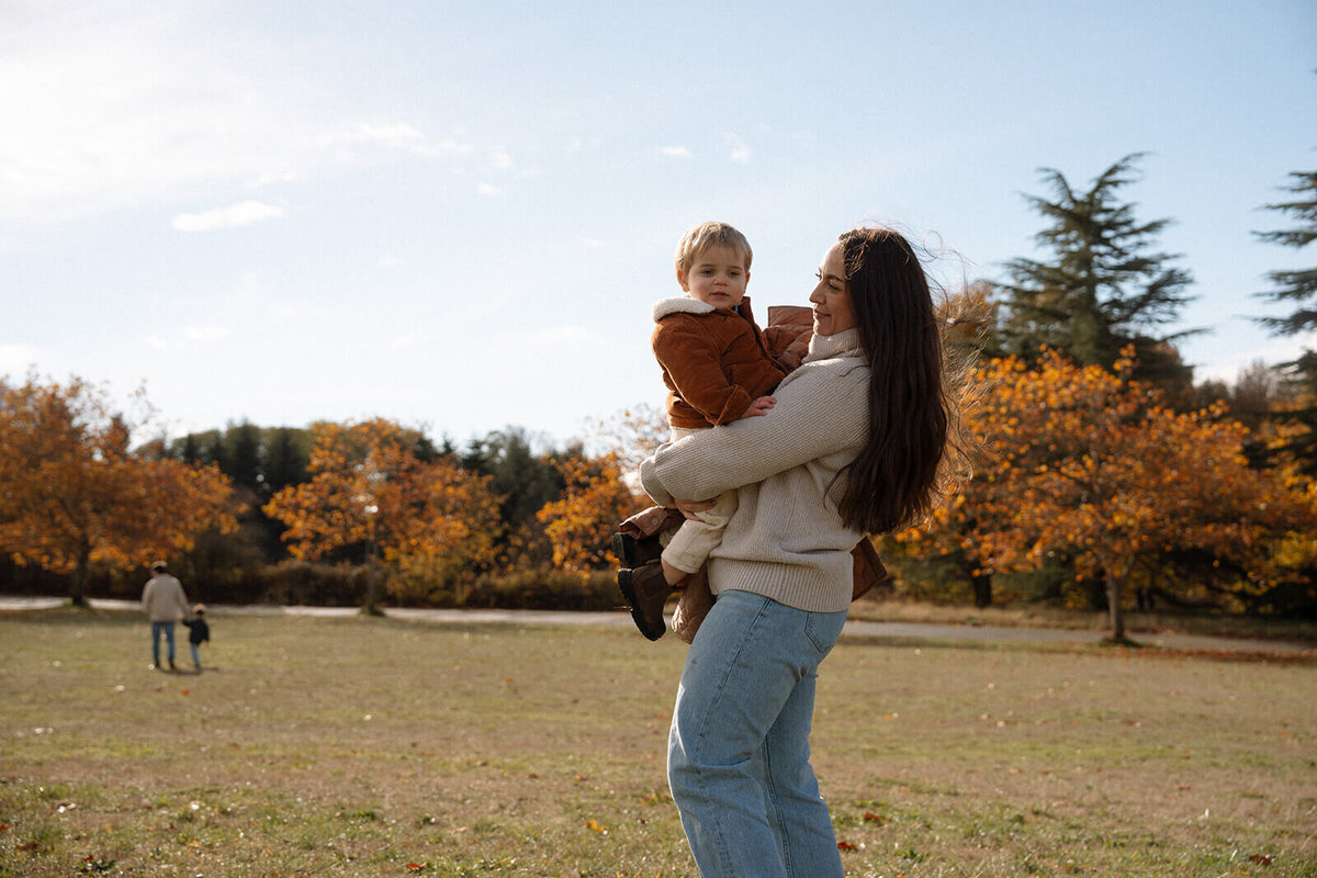 mother holding son in park with dad in background at park in fall