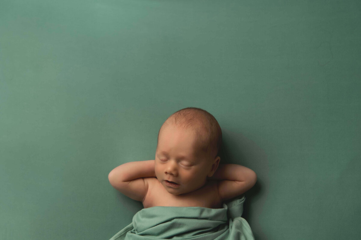 A newborn photograph by Mobile, Al Newborn Photographer of a baby boy sleeping peacefully on a sage green backdrop with his hands tucked comfortably behind his hand with a matching sage green swaddles draped over his body for warmth.