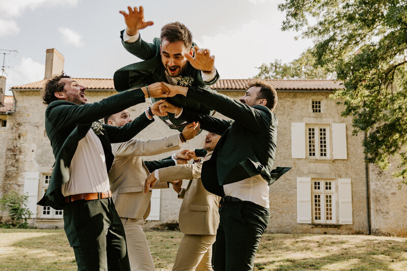 Marié jeté en l'air par ses amis devant une vieille bâtisse en pierre pour une séance photo mariage en vendée.