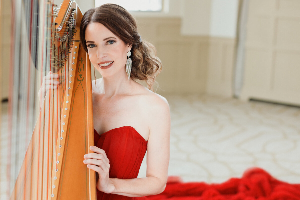 light skinned woman with dark hair in red gown that is draped on the floor behind her. Gold harp is leaning on her shoulder as she looks at camera and smiles.