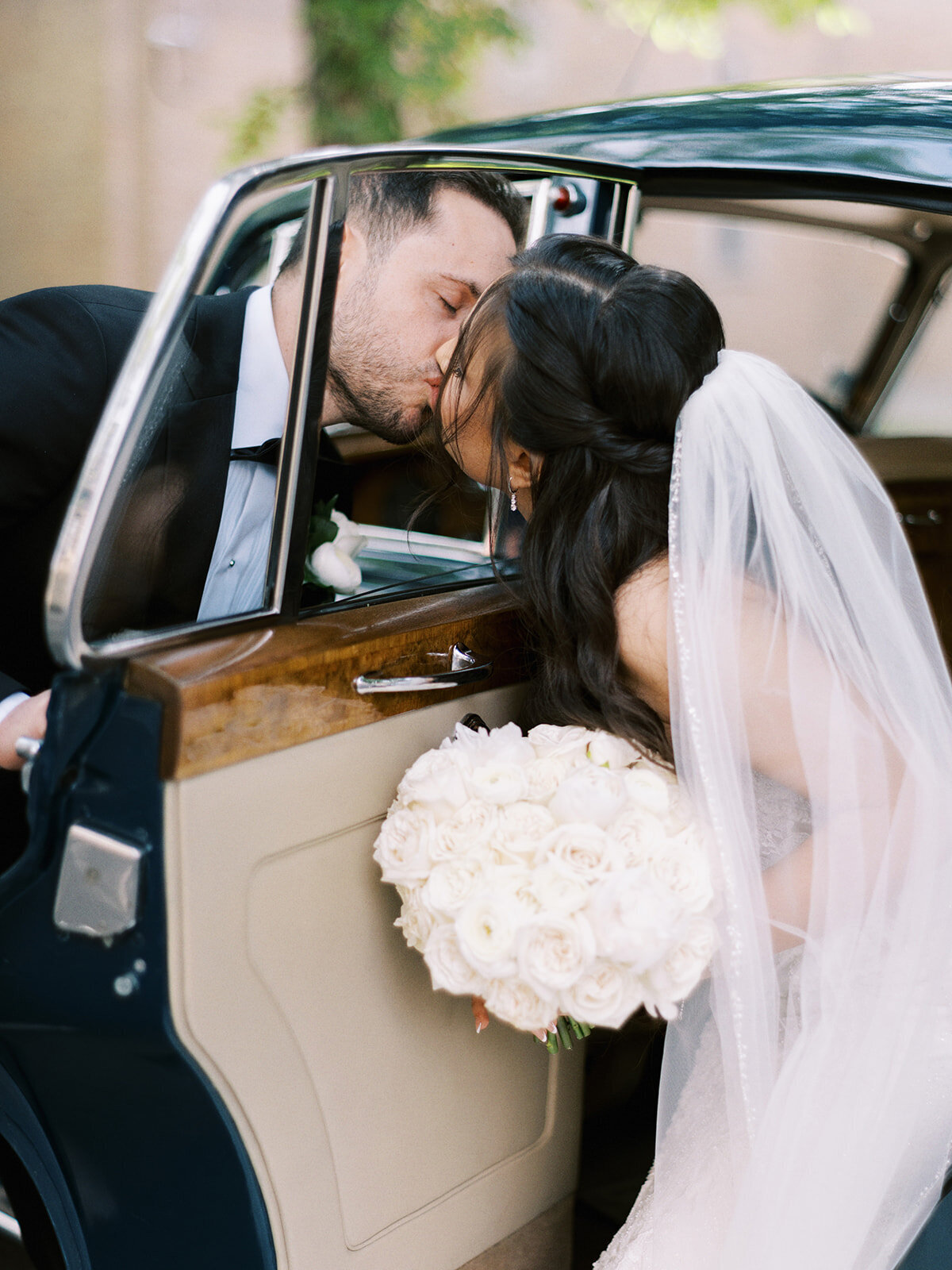 A newlywed couple shares a kiss in front of a vintage car. The bride holds a bouquet of white roses, and the groom wears a black suit with a boutonnière, capturing the elegance of a classic Calgary wedding.