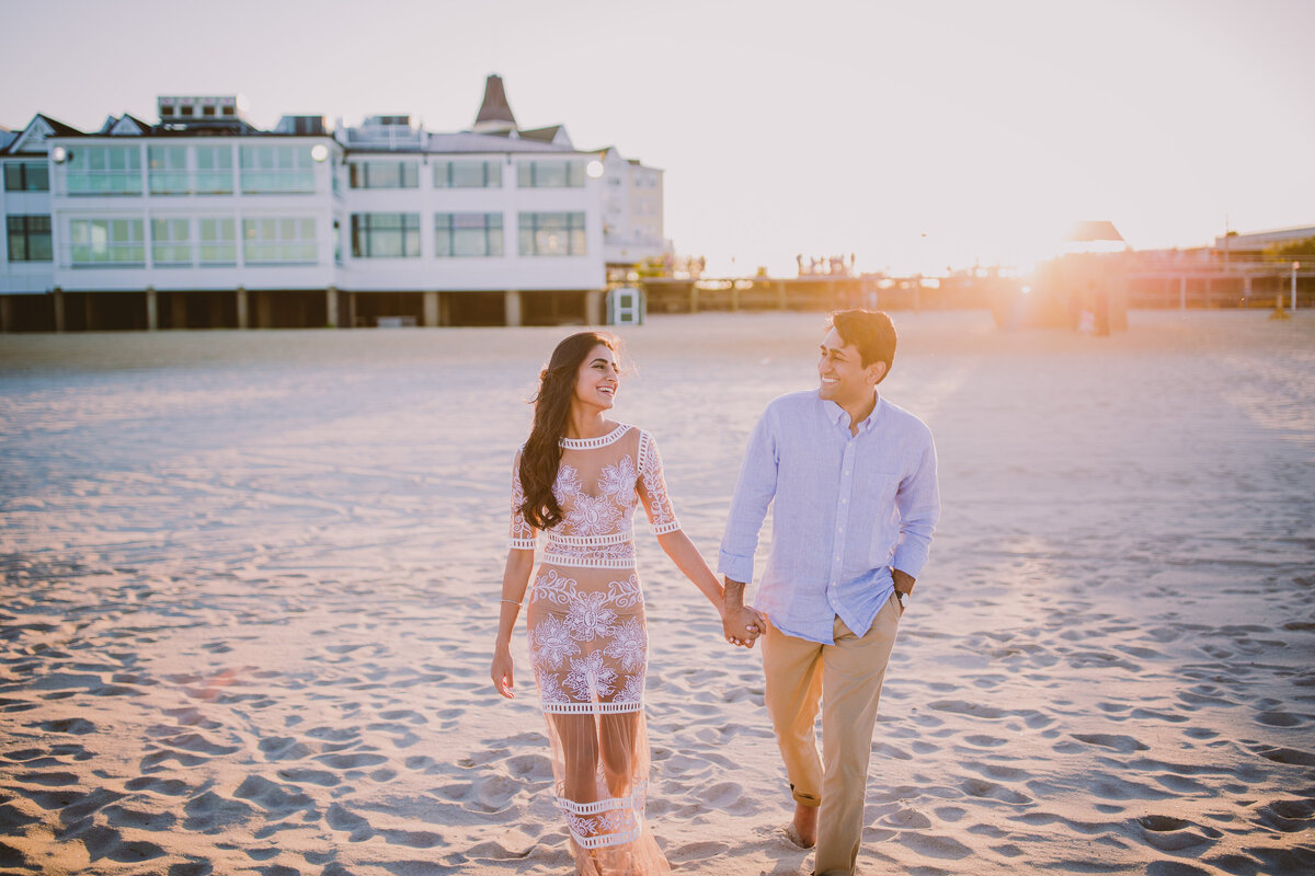 Engaged couple walking on the beach smiling at each other with the sun behind them at long branch beach in nj