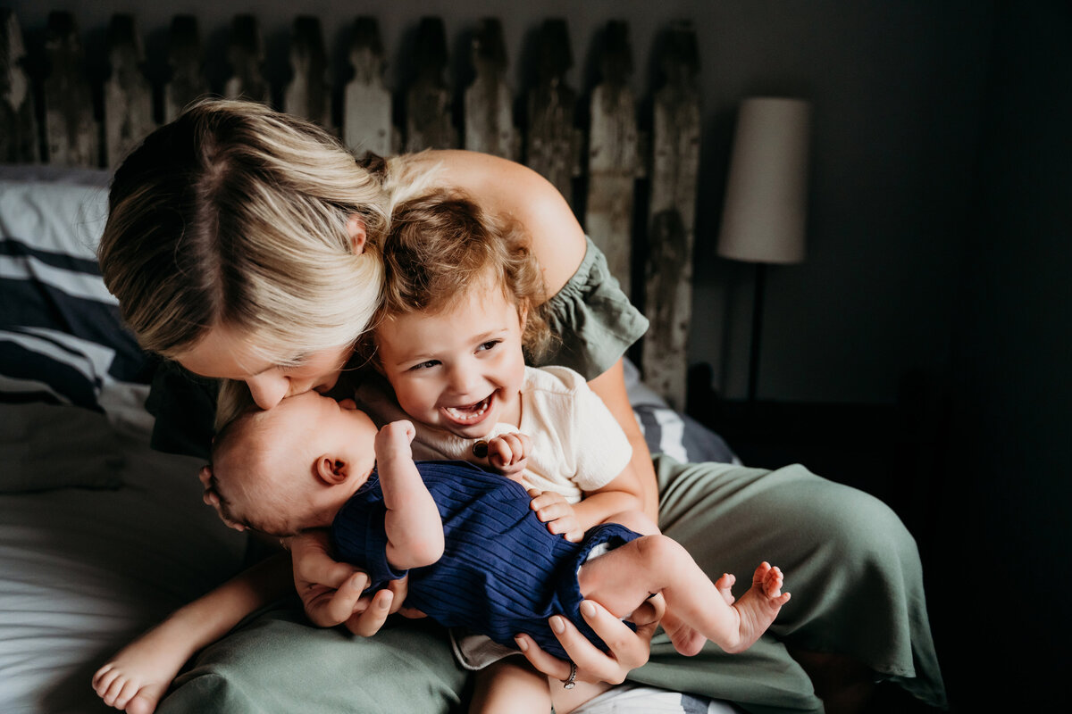Newborn Photographer, a mother kisses her baby as she holds him and her older sister on the bed