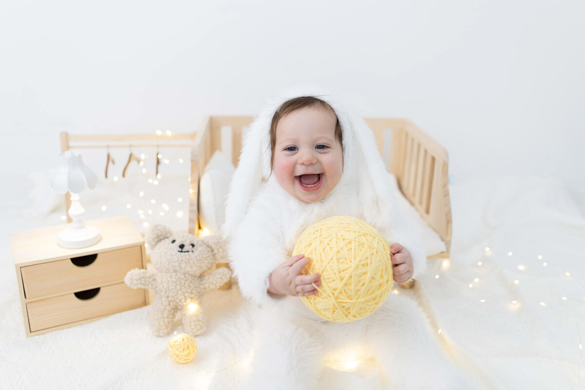 Baby in white fuzzy outfit sitting on mini bed surrounded by fairy lights - winter-themed milestone session
