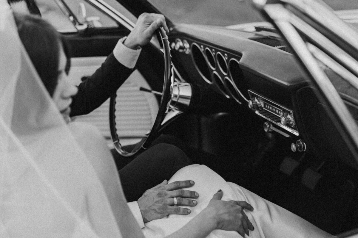 Black and white image of bride in gown holding steering wheel of vintage car.