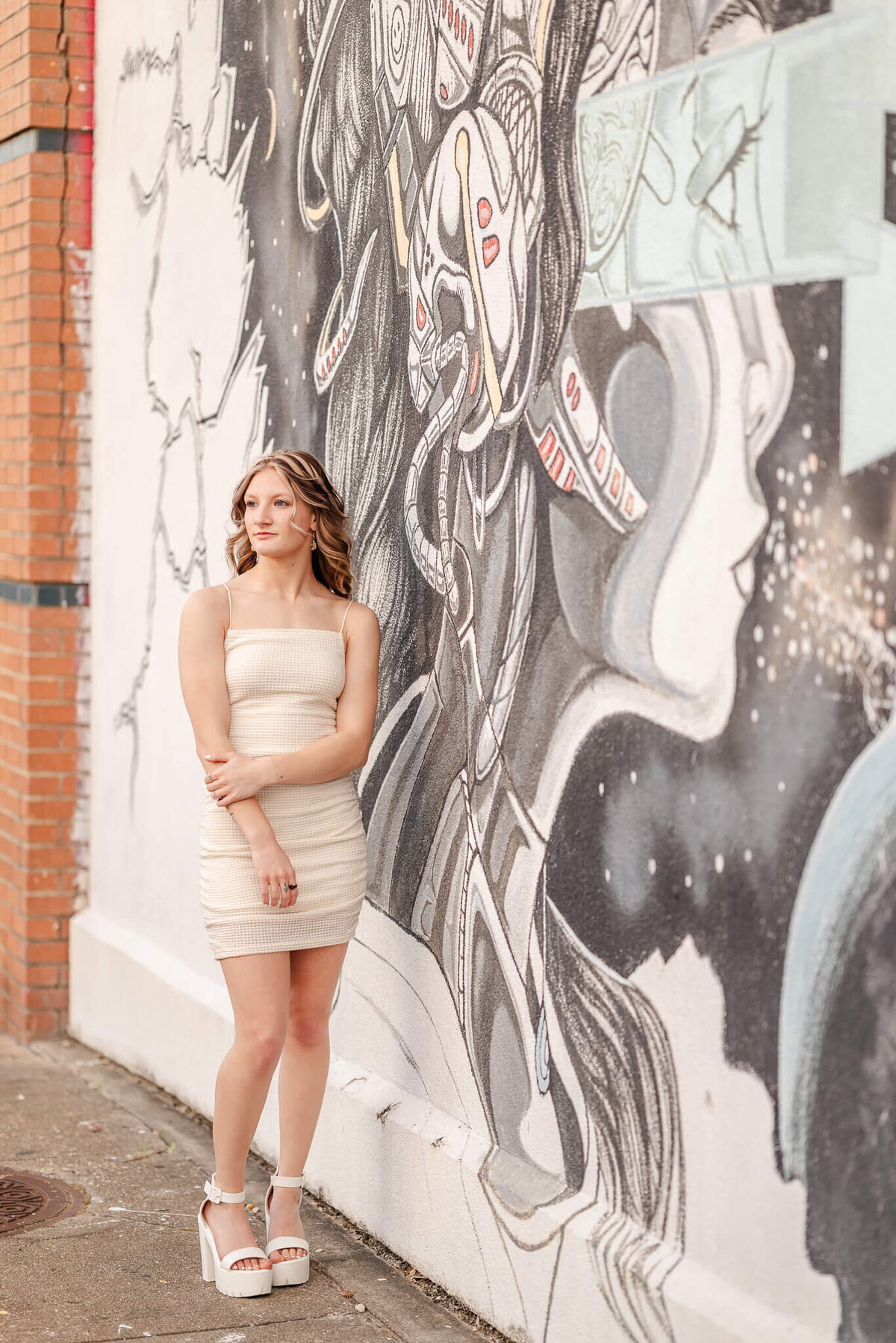 A high school senior, wearing an off-white bodycon dress and shoes, stands by a black and white mural in the Norfolk Neon District.