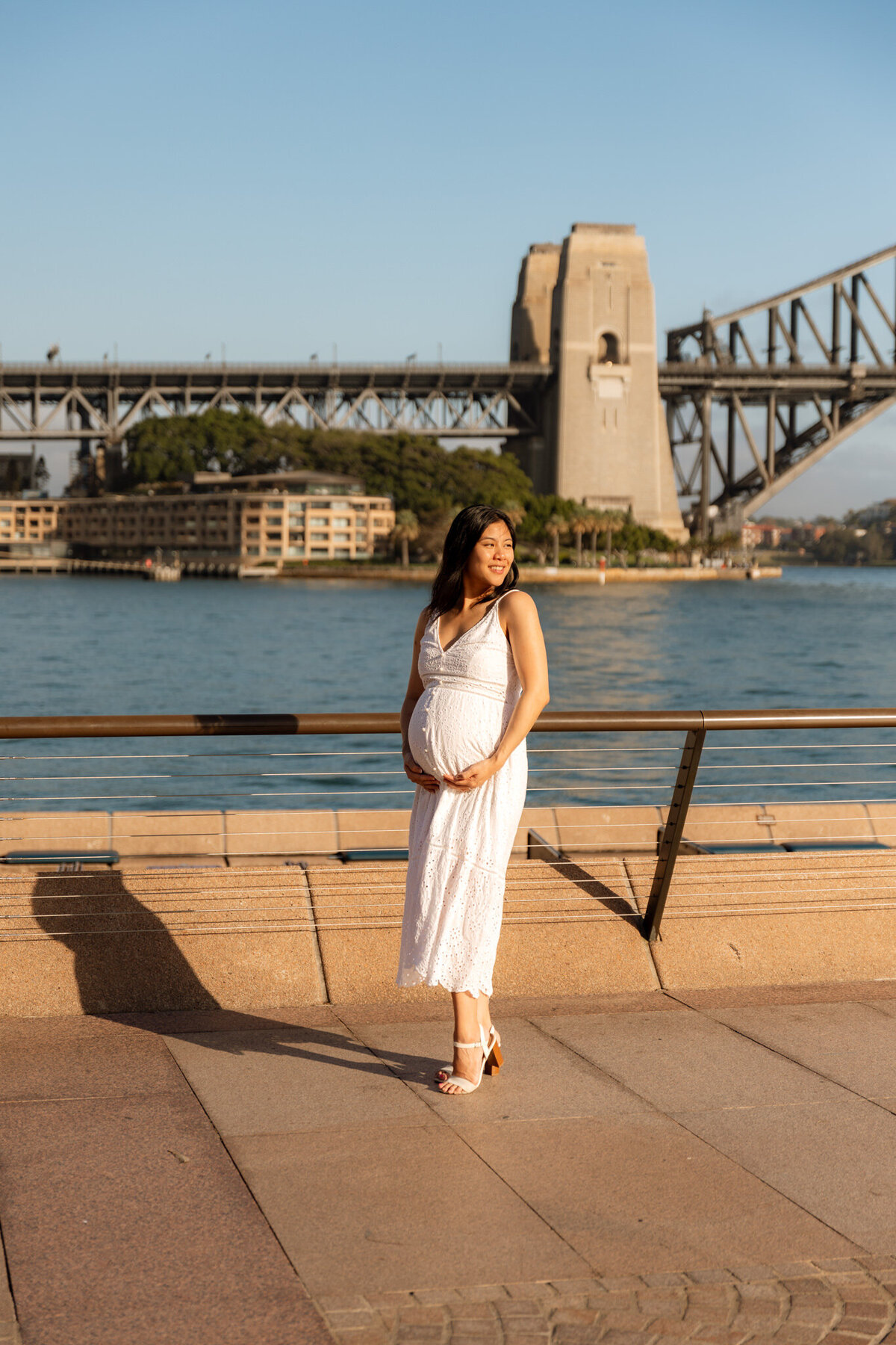 Pregnant woman in white dress standing near Sydney Harbour with Harbour Bridge in the background during golden hour maternity session