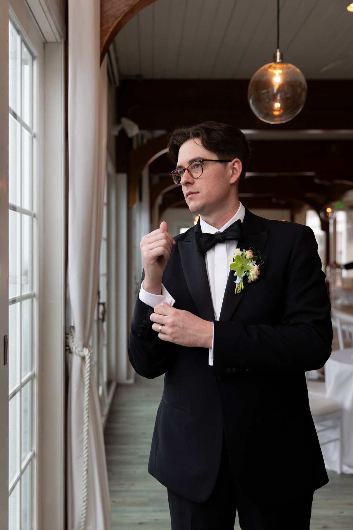Rhode Island Wedding Photographers | Kelsey Sheehan Photography Timeless Rhode Island Weddings | A man in a tuxedo stands by large windows, adjusting his cufflink. He wears glasses and has a floral boutonniere. The setting is elegant and formal.