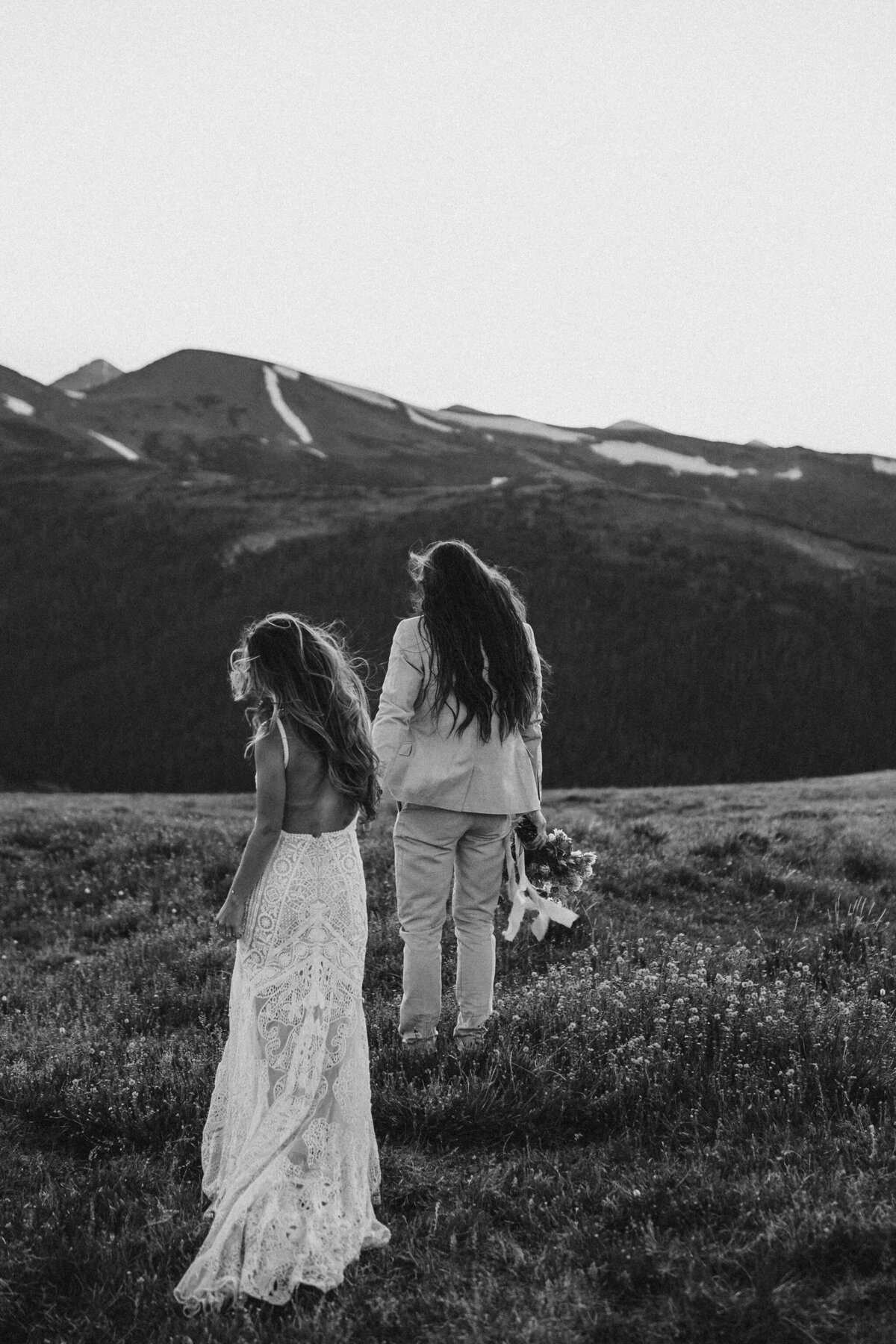 bride and groom wearing a wedding gown and suit walking in the mountains while groom holds a bouquet