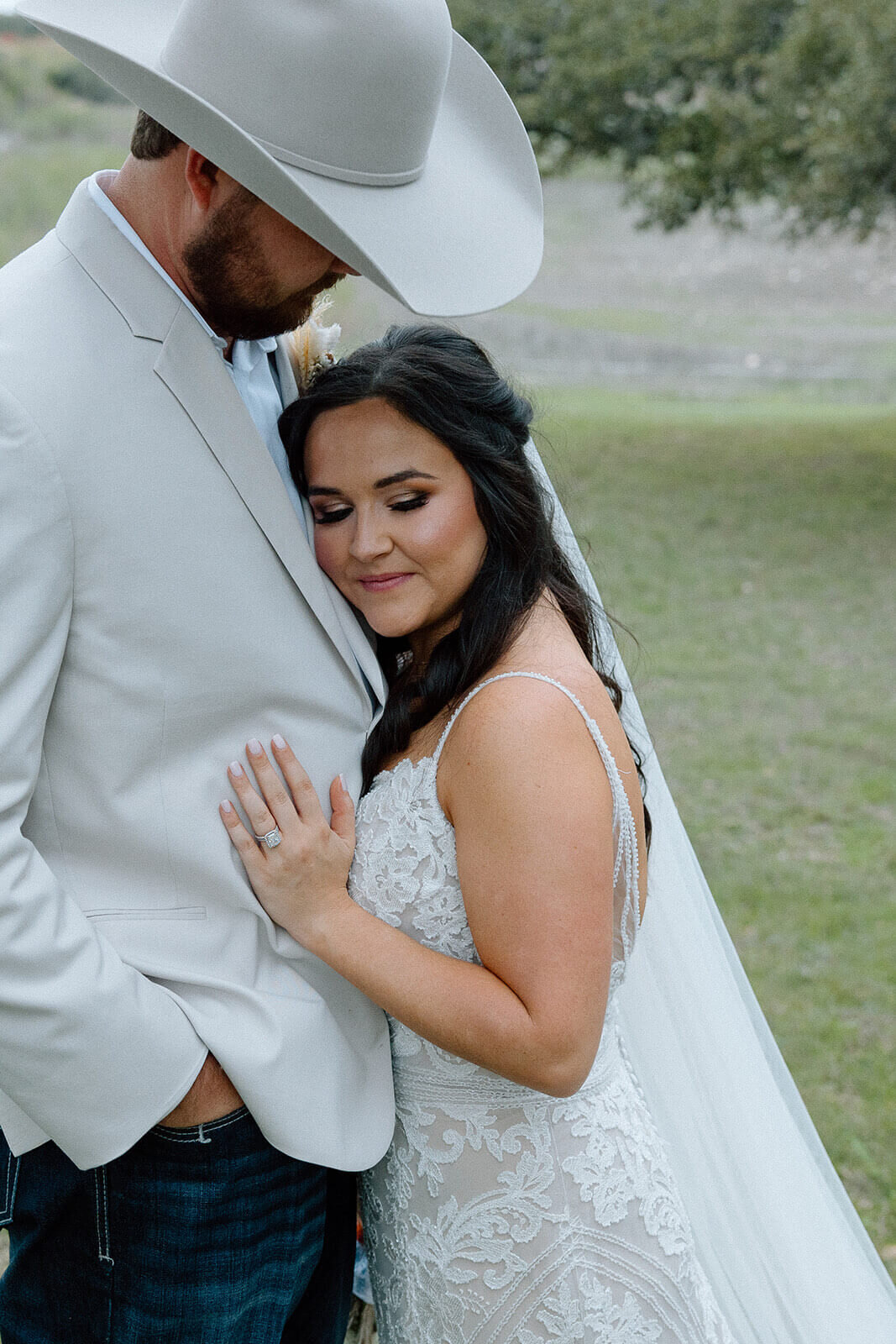 A glowing bride in a lace wedding dress embraces her groom in a soft beige suit and cowboy hat during their Texas hill country wedding. Her bridal glam features natural tones, a soft smokey eye, and flawless airbrush foundation. Mistique Makeup offers professional mobile bridal hair and makeup in San Antonio, Austin, Dallas, and nearby ranch venues. Perfect for brides seeking a timeless, elegant look with a touch of rustic romance.