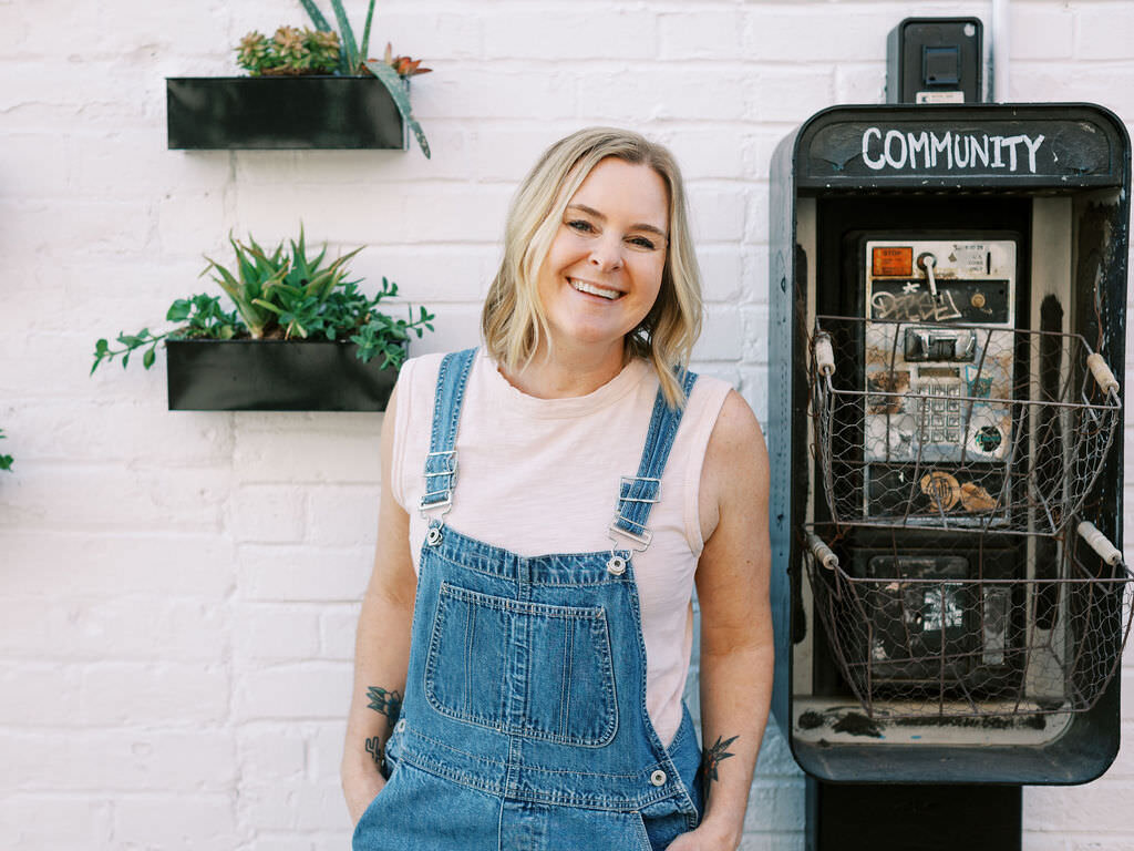 A woman in overalls smiling while standing next to an old payphone.