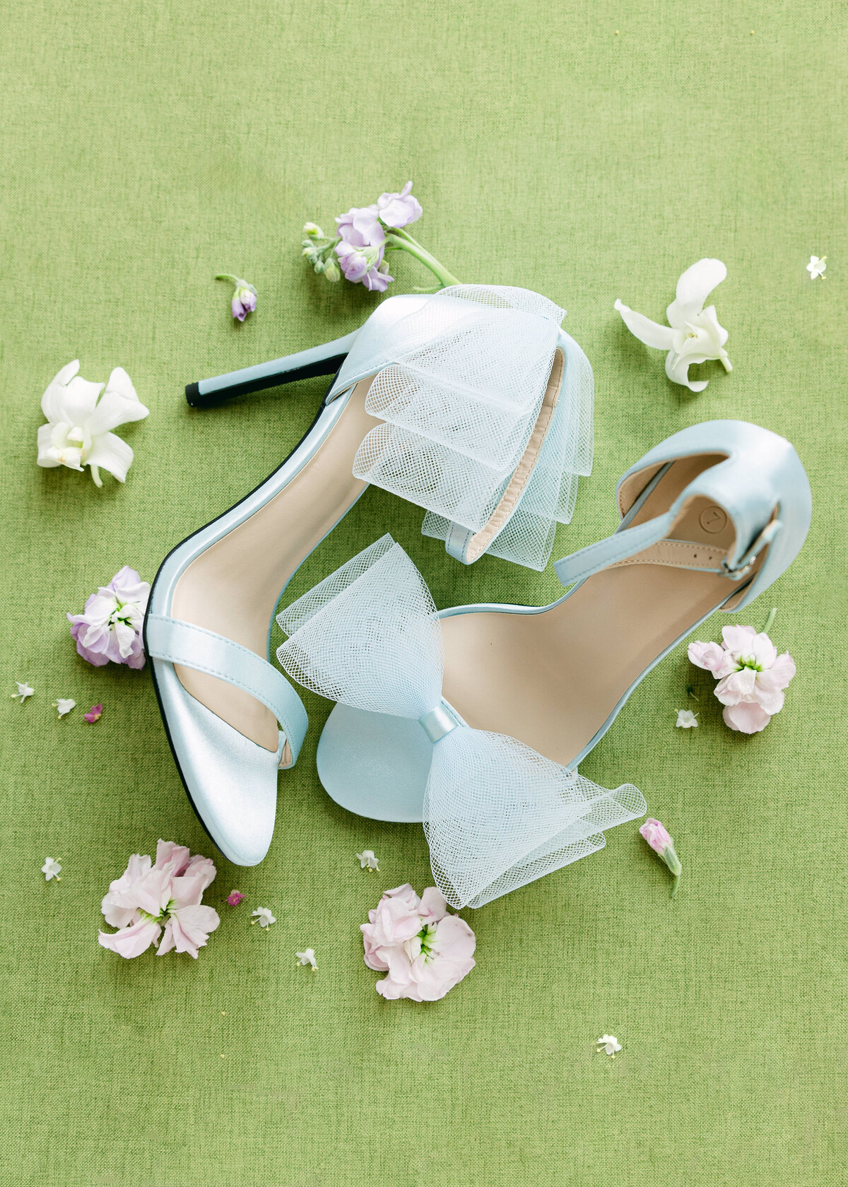 Powder-blue bridal heels with tulle bows styled on a fresh green backdrop with scattered pastel blooms—detail from a Savannah wedding, captured by luxury wedding photographer Amia Marcell.