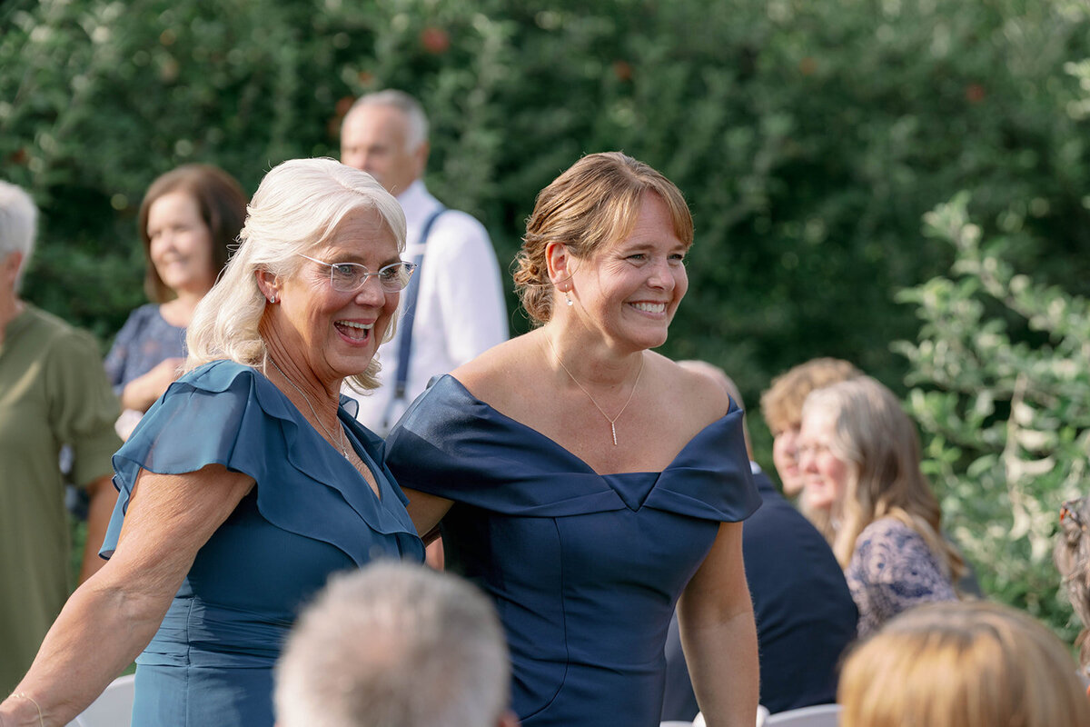 The mother of the bride and mother of the groom walking together and laughing as they exit the ceremony at a September orchard wedding in Frankfort MI.