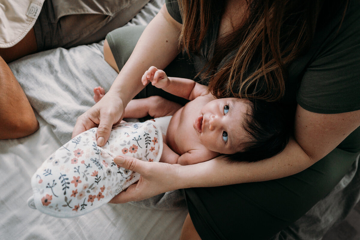 a dark haired dark eyed newborn looks up at his mother wide eyed.  She is cradling him between her legs and arms but her face is not visible. She is also holding a floral fabric.