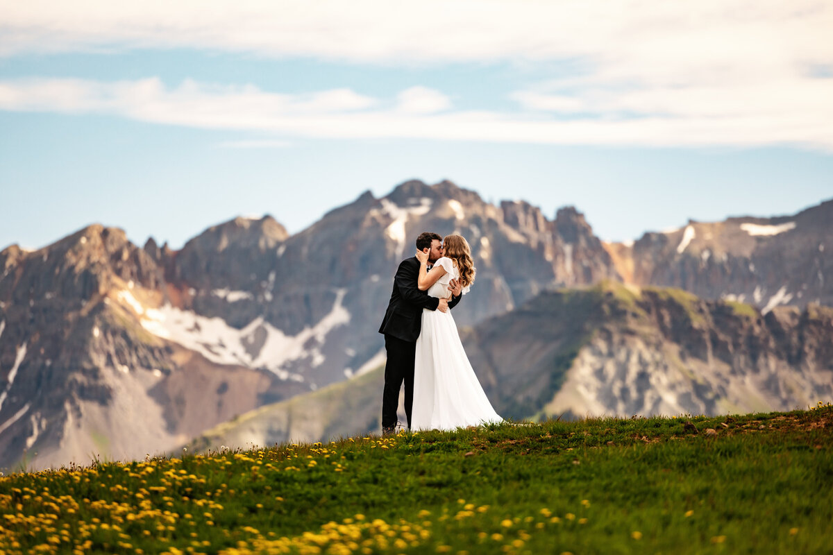 Shannon Lee, from Love Like Wild Photography, captures a couple embracing on a grassy hill with majestic mountains in the background.