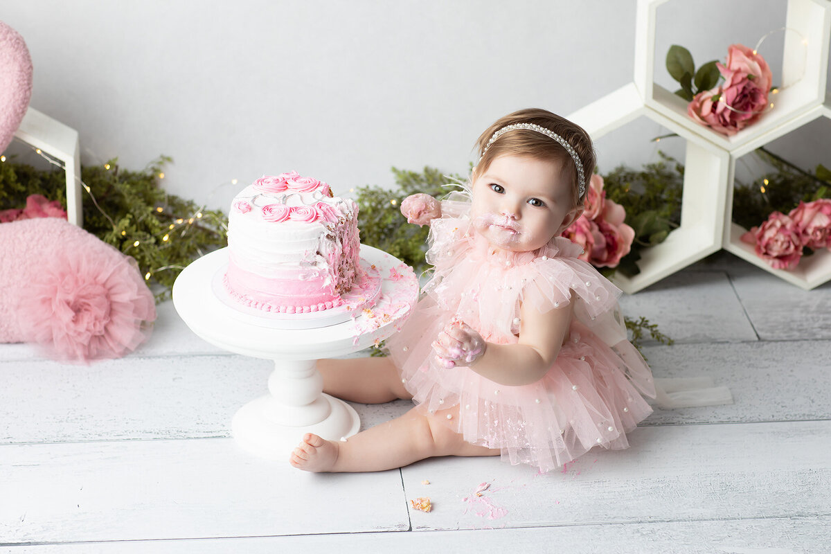 DFW milestone photographer captures a baby girl in a ruffled pink tulle dress enjoying a cake smash session with a half-eaten pink and white cake, frosting on her face and hands, surrounded by floral and soft décor.