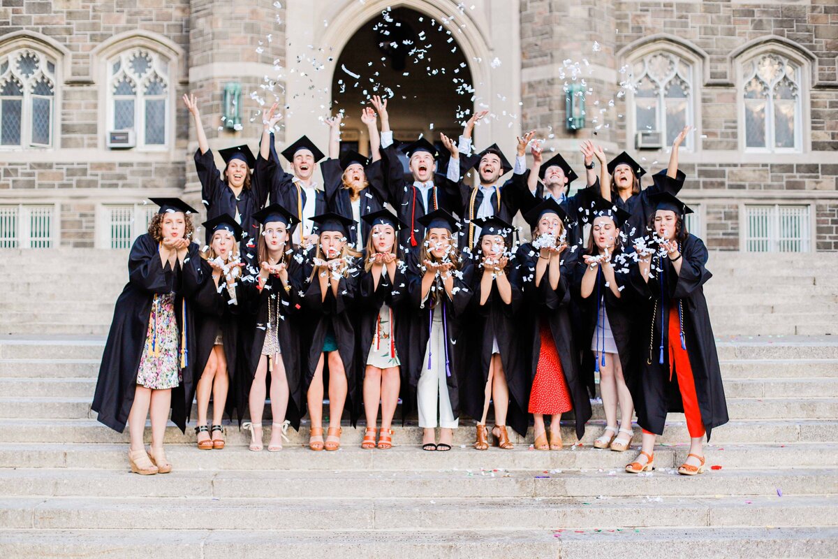 Large group photo of girls in caps and gowns blowing confetti on a staircase