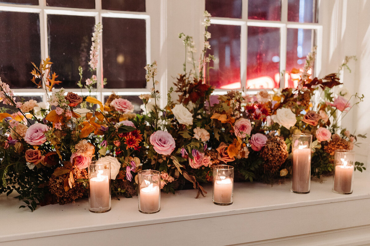 Elegant window arrangements and candle moments create a warm autumnal setting in hues of dusty rose, burgundy, taupe, mauve, and lavender florals composed of roses, raintree pods, ranunculus, spray roses, copper beech, and fall foliage. Design by Rosemary and Finch in Nashville, TN.