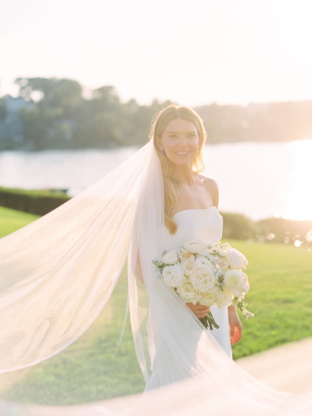 Wedding Photographer RI | A bride in a white gown and veil stands outdoors, holding a bouquet of white flowers. The sunset creates a soft, warm glow