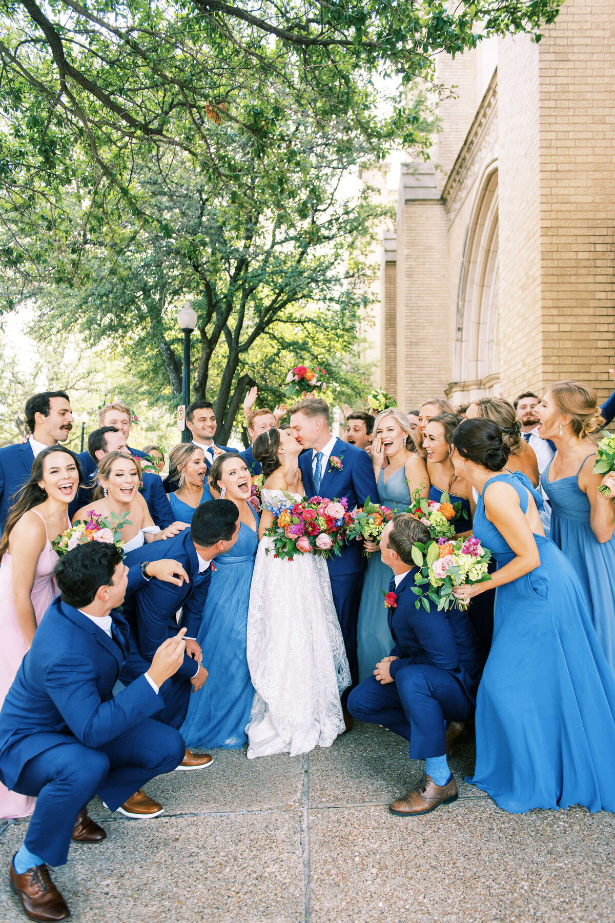 Large wedding party in blue celebrates as bride and groom kiss outside of The 4Eleven venue in Fort Worth