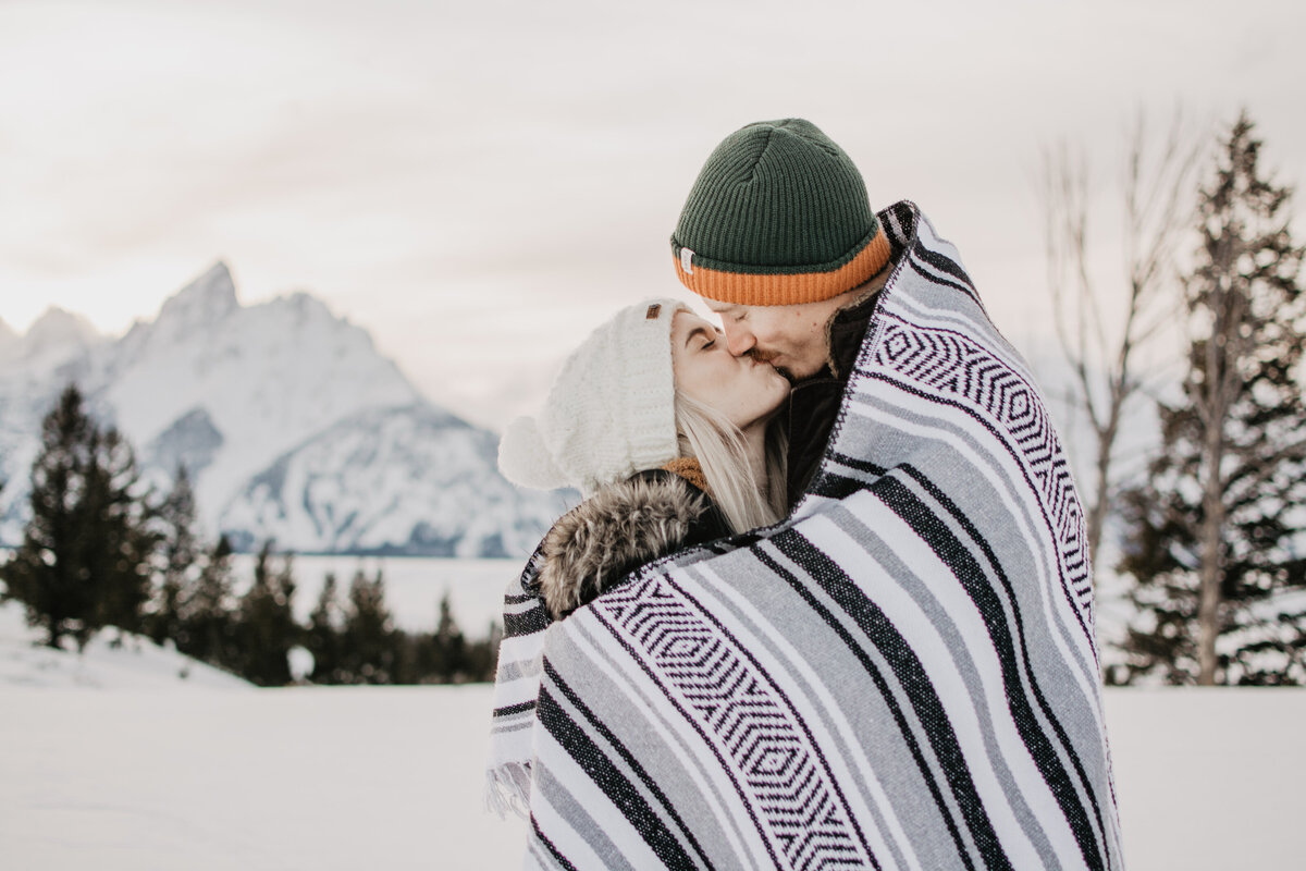 jackson hole photographers captures young couple kissing as they are wrapped up in a boho aztec blanket  in Jackson Hole for a winter engagement session