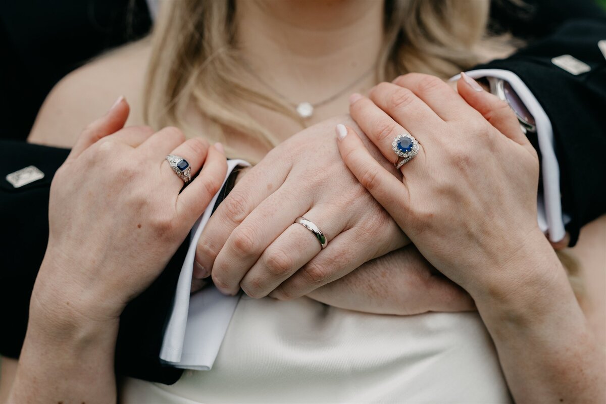 A close photograph of two newlyweds linking hands. We see their wedding rings in clear view.