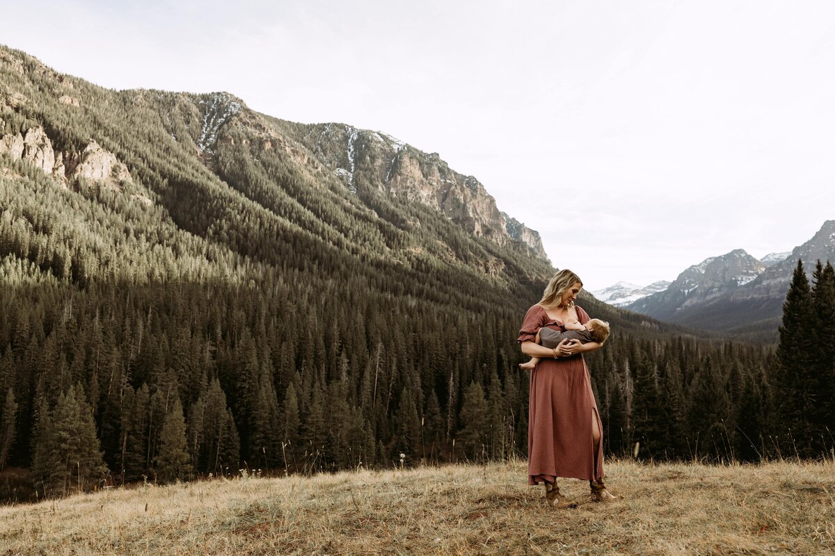 Breastfeeding mom holding her baby while smiling and looking happy.  Background is the mountains.