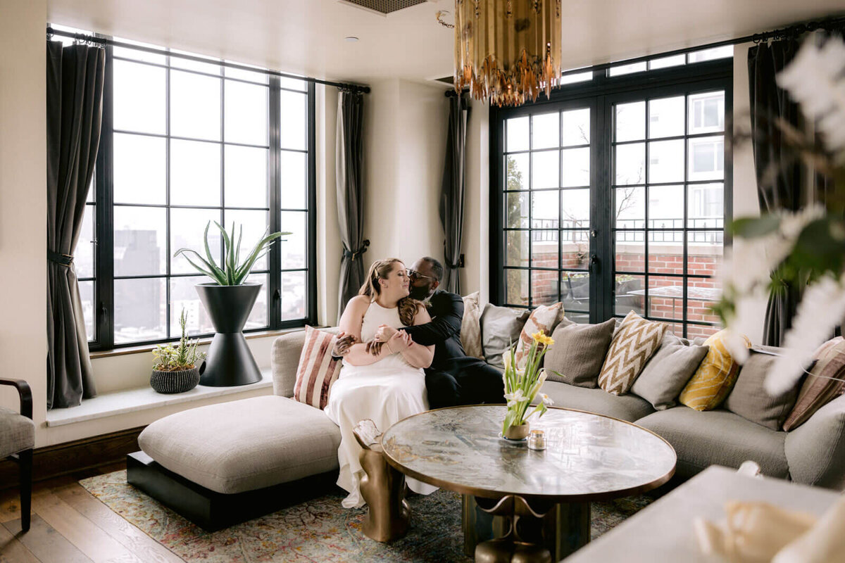 The groom is kissing the bride while sitting on a couch inside a Ludlow Hotel room.