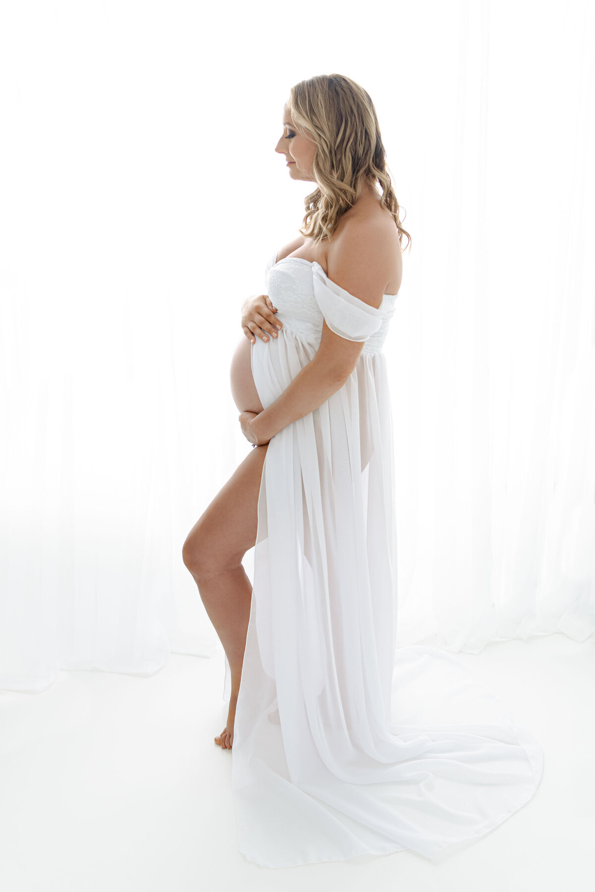 Backlit image of expecting mom holding belly dressed in white sheer gown