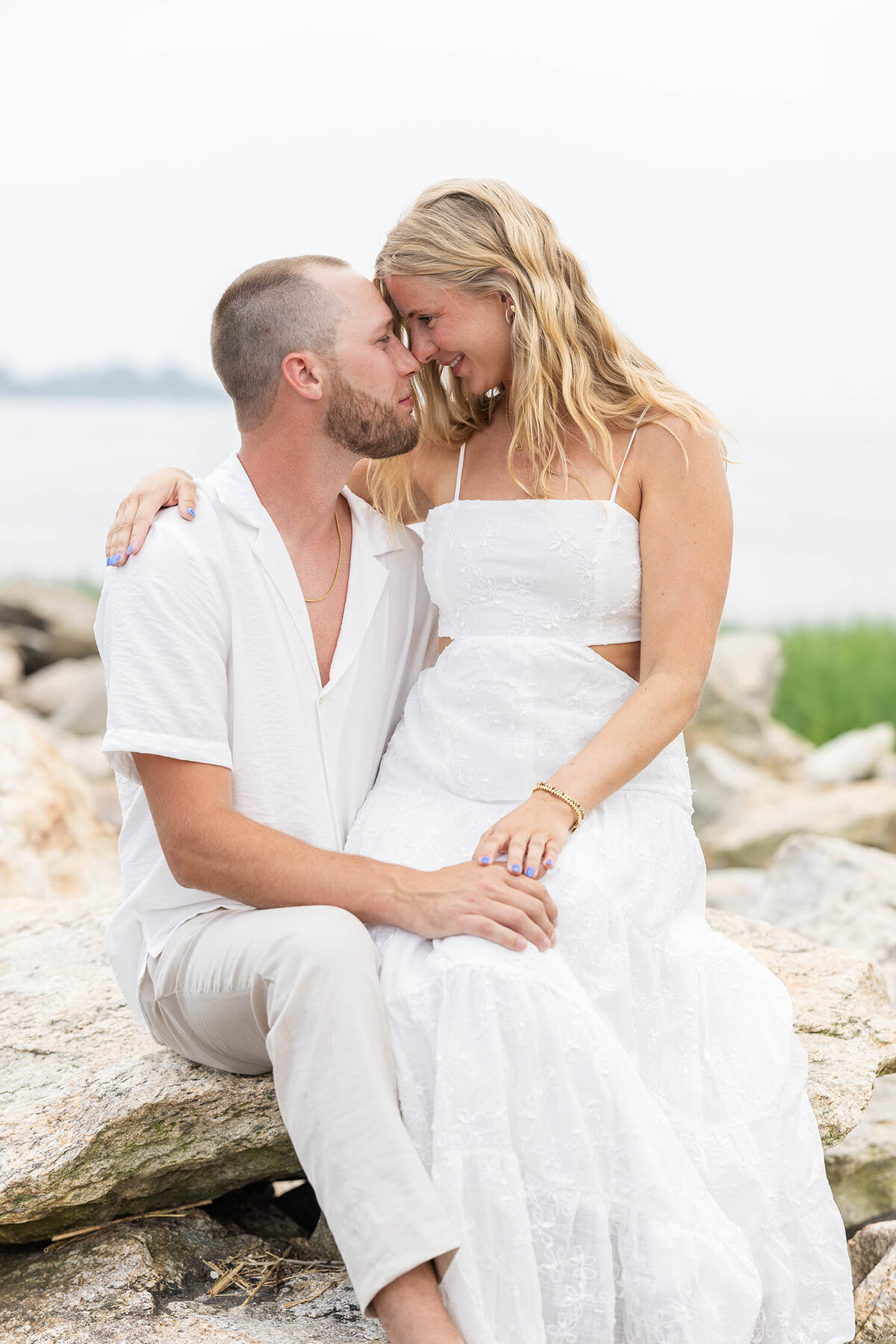 Couple sitting on a rock at a rocky shoreline during a romantic Coastal Connecticut Session, looking at each other with both their nose touching
