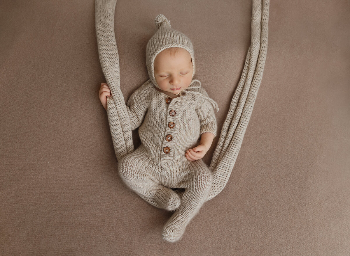 Aerial image of a newborn photoshoot. Baby is wearing a beige knit romper and hat. There is fabric draped under him to give the illusion that he is sitting on a swing. Captured by top Brooklyn newborn photographer Chaya Bornstein