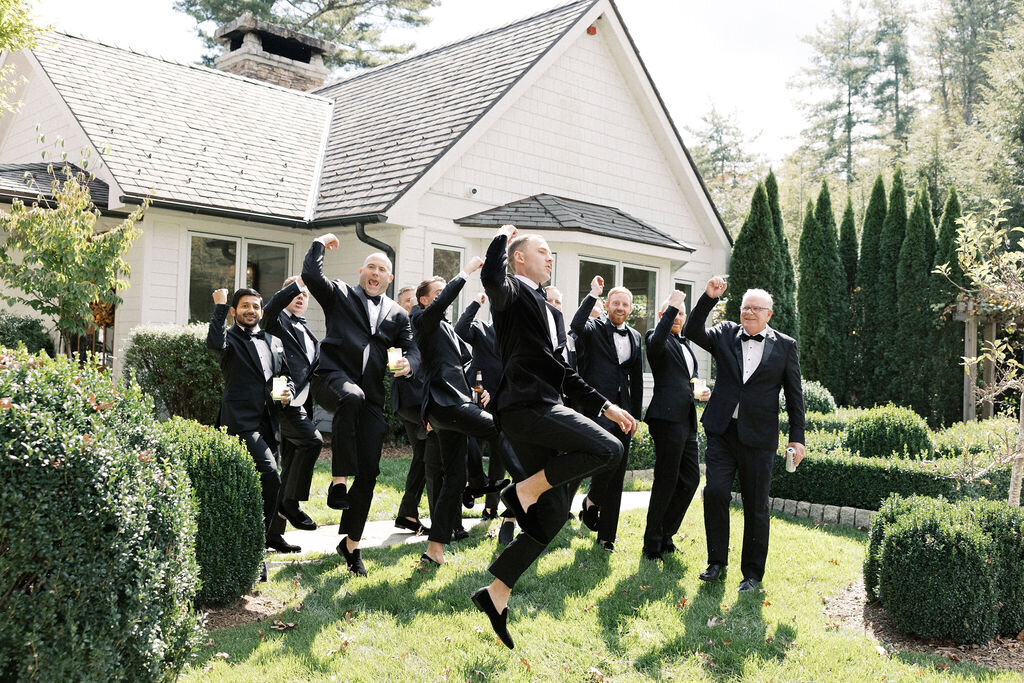 Groomsmen in tuxedos jumping and celebrating outside the wedding venue.