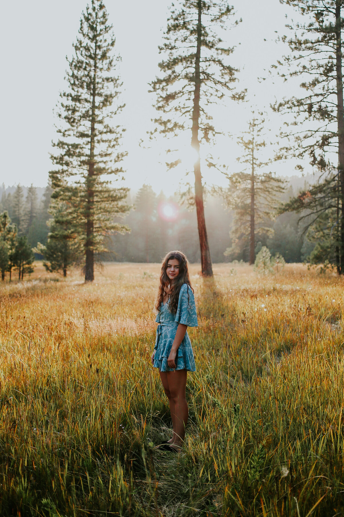 Young woman stand in golden forest meadow wearing short green jumper as sun sets.