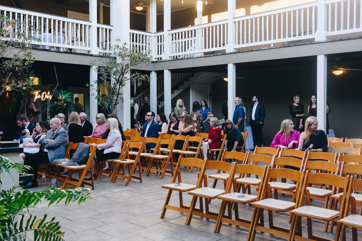 A group of people sit and stand in a courtyard with rows of empty wooden chairs, preparing for an event. Some guests chat while others wait. The setting is bright, with balconies and plants visible.
