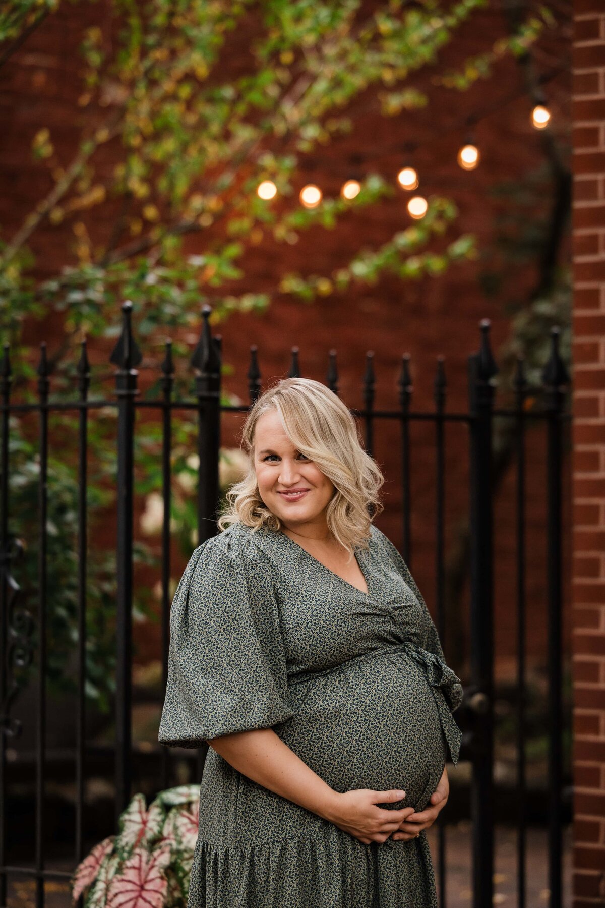 Pregnant woman smiling outdoors with a hand on her belly, captured by a maternity photographer in Pittsburgh, standing by a black fence with string lights in the background.