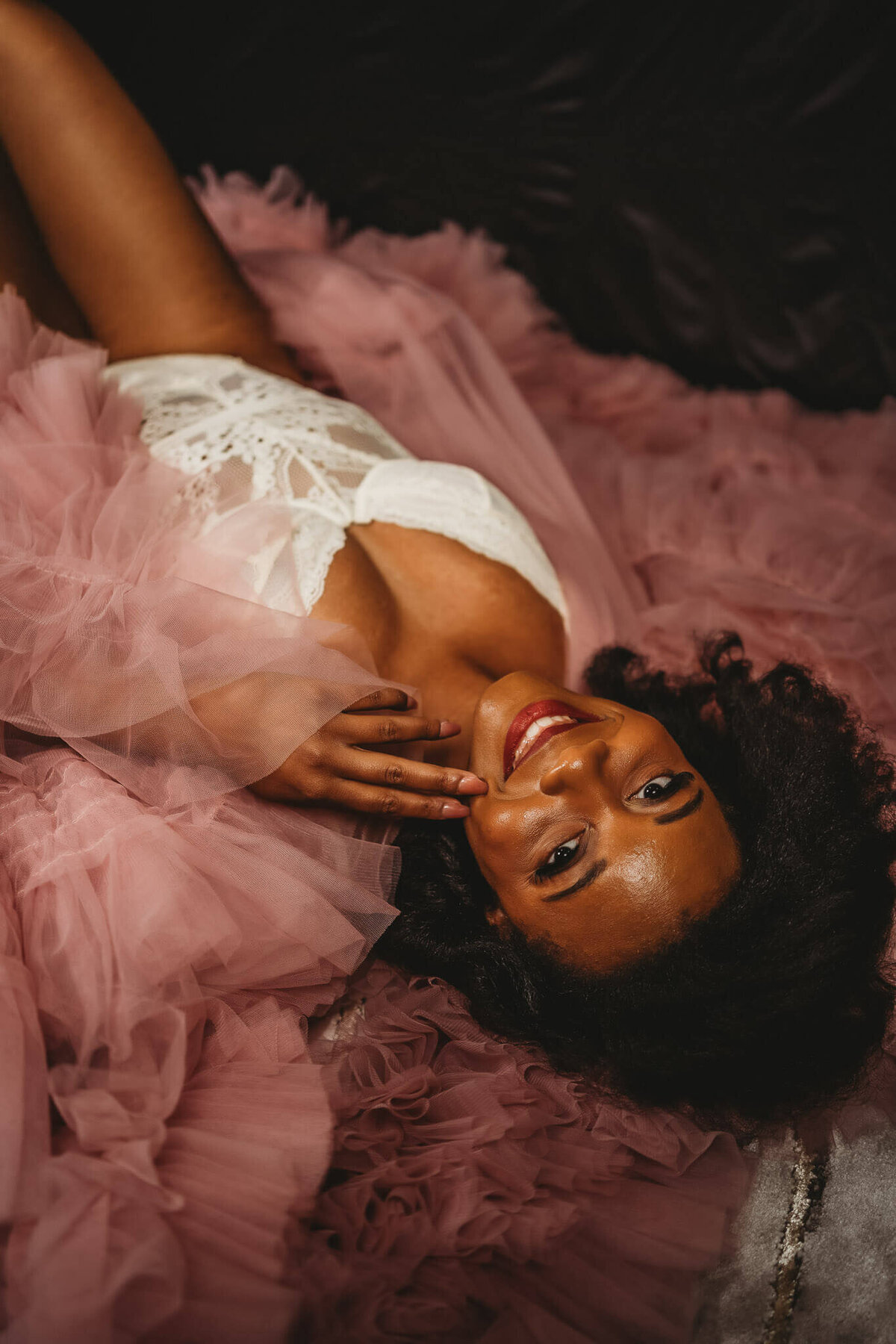 beautiful black woman laying on giant pink tulle robe, smiling at camera with her hand on her face