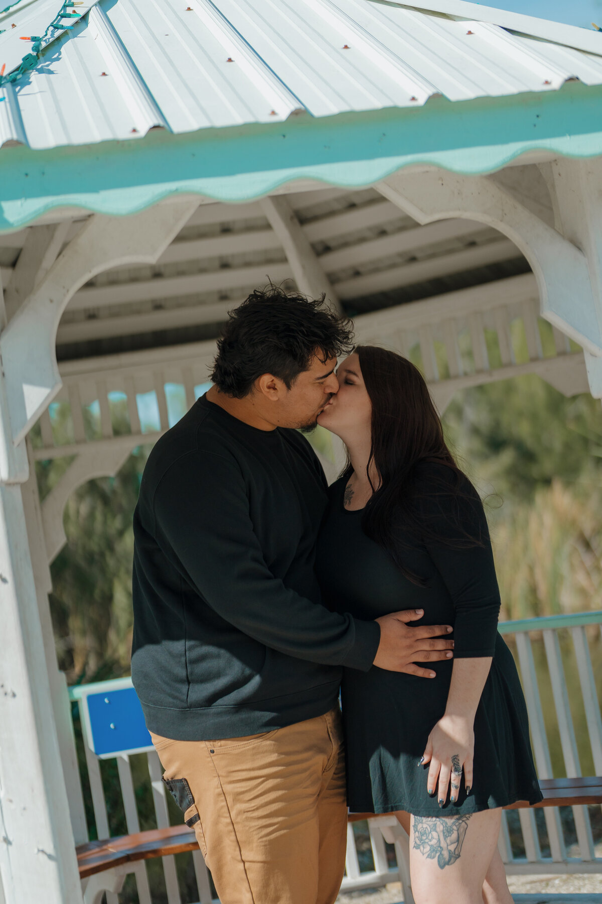 Couple sharing a kiss under a gazebo with a white roof, surrounded by greenery
