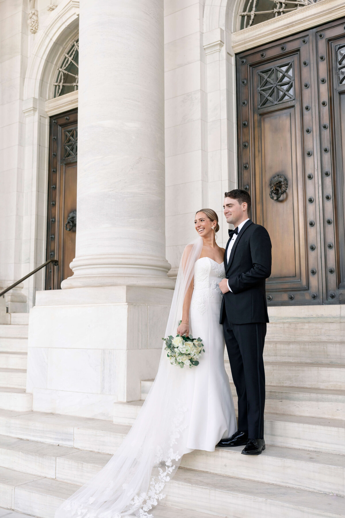 A bride and groom stand on white marble steps in front of large wooden doors. The bride wears a white gown with a long train and holds a bouquet. The groom is in a black tuxedo. Both look off to the side, smiling.