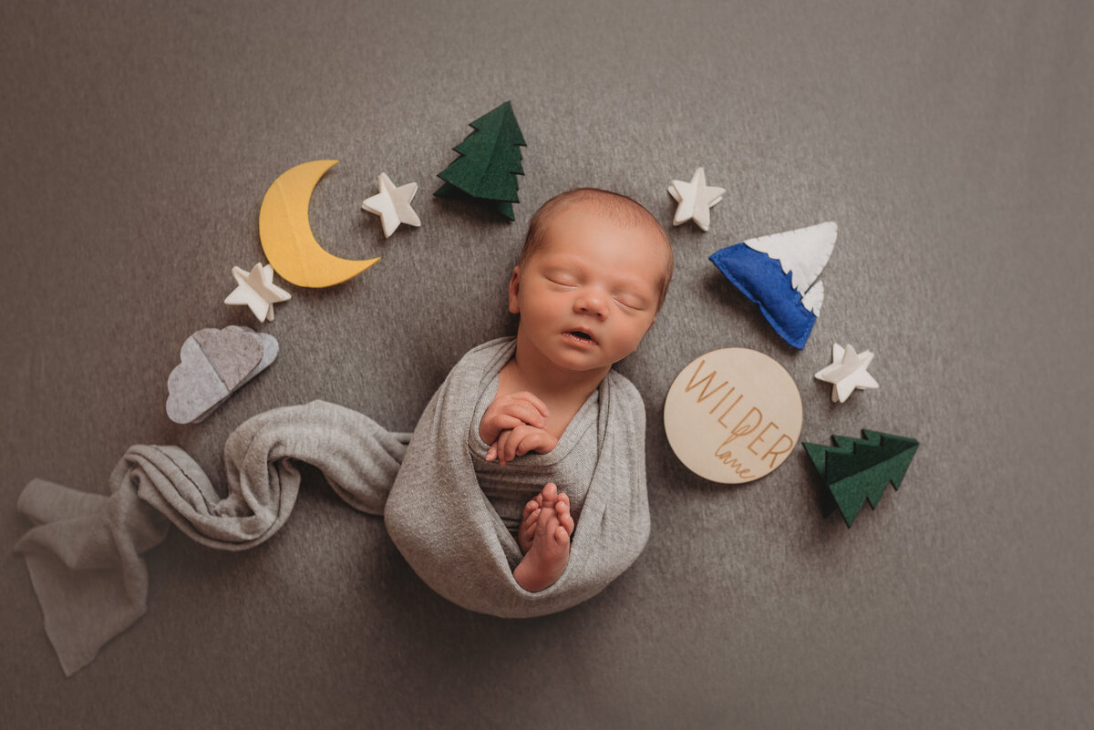 Newborn boy asleep wrapped in gray swaddle on gray fabric backdrop with moon, stars, pine trees and mountain props around him